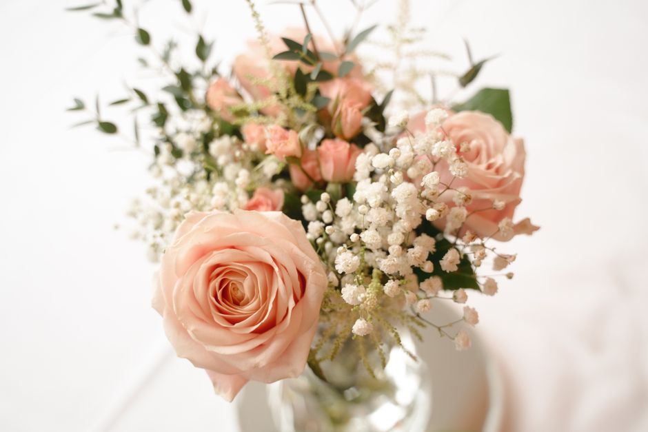 A floral arrangement in a clear glass vase featuring a mix of soft pink roses with tightly wound petals and gently unfolding blooms, accented by clusters of delicate white baby's breath and sprigs of 