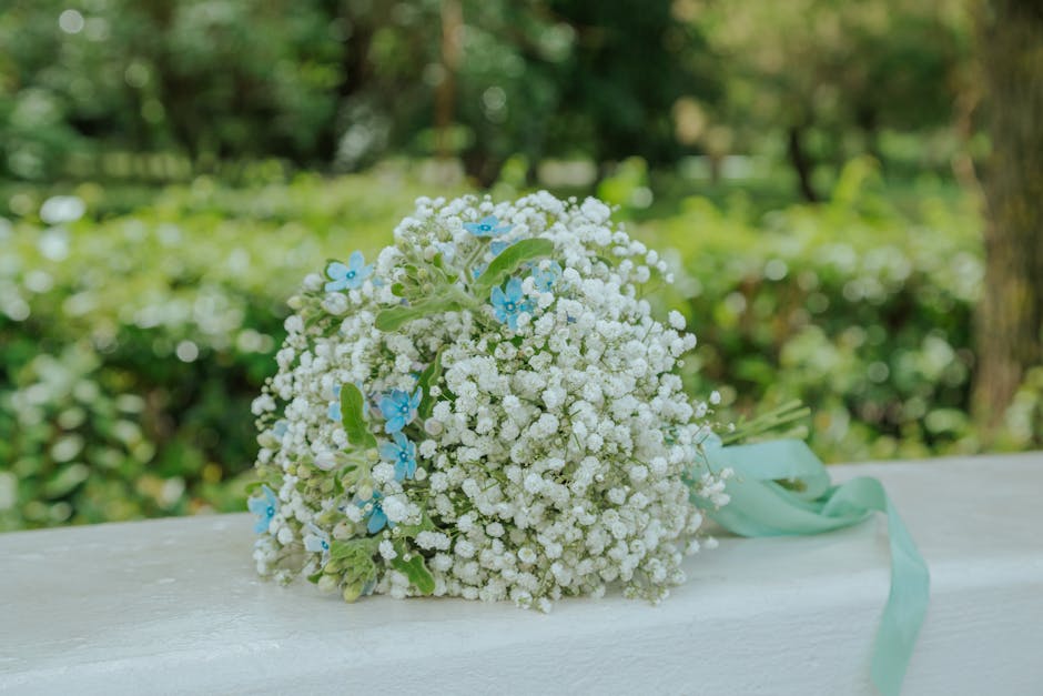 A bouquet of fresh white baby's breath flowers with small, delicate clustered blooms, accented by a few light blue flowers and green leaves, tied together with a pale mint-green ribbon. The bouquet is