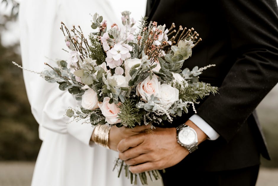 A close-up image of a couple holding a delicate bouquet of fresh flowers, featuring pale pink roses, white hydrangeas, and soft green eucalyptus leaves arranged in a loose, natural style. The bouquet