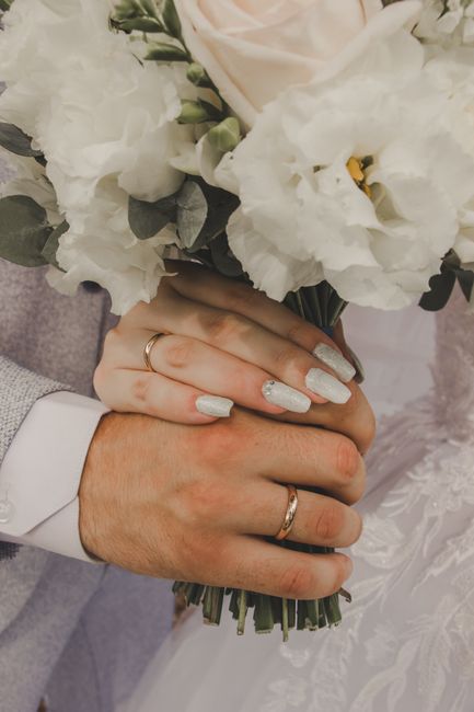 Close-up of a bride and groom holding a bouquet of fresh, predominantly white flowers, including eustoma and lisianthus, with hints of green foliage. The bouquet is wrapped with a simple, natural-colo
