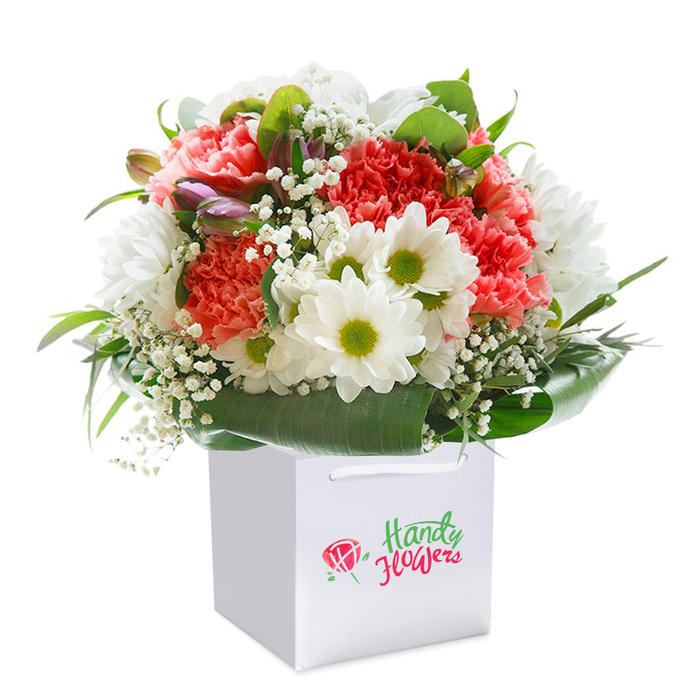 In the heart of Highams Park, this vibrant bouquet evokes a sense of seasonal delight, presented in a spotless white square gift bag with tidy ribbon handles ready for gifting or delivery. The arrangement bursts with coral and peach carnations, their abundant ruffled petals inviting the gaze and conjuring softness reminiscent of early spring mornings in Vincent Road gardens. Creamy white daisies, each sporting a lively burst of yellow-green at the centre, mingle playfully among the larger blossoms, adding notes of cheerful brightness and calm. Wisps of baby's breath thread between the main flowers, forming delicate, cloudlike bridges, while unopened purple-tinged buds nestled to one side hint at new beginnings and the ever-renewing cycle of nature. The bouquet is encircled by substantial, lustrous deep green leaves that cradle and frame the whole creation, drawing inspiration from the lush parkland that gives the area its name. Lit by a gentle, even light that highlights texture and saturation, the finely rounded shape and considered placement of every stem and bloom celebrate both freshness and expertise. Designed to uplift and inspire-whether as a birthday gift, surprise celebration, or just a thoughtful touch-this bouquet is a testament to the skill of Highams Park's local florists, bringing a tailored experience to every heartfelt moment.