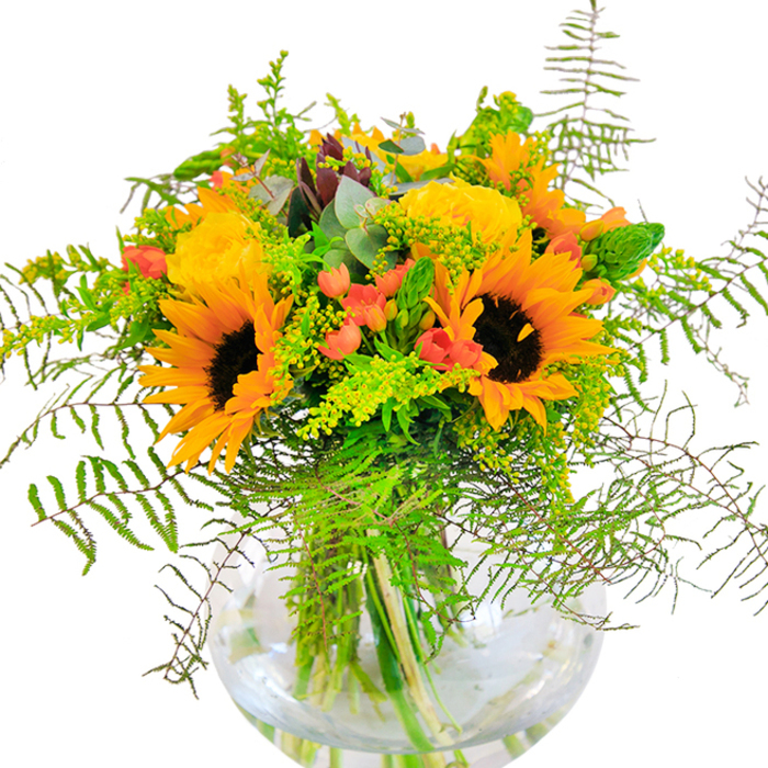 With a radiant blend of yellows, oranges, and pops of red, this bouquet delivers an immediate sense of warmth and exuberance-much like a summer afternoon in Highams Park. Sturdy sunflowers, with their wide, velvety petals and deep brown-gold eyes, create a glowing focal point, flanked by golden garden roses whose tightly packed petals spiral outward with subtle elegance. Glossy crimson Hypericum berries nestle between the blooms, their vibrant sheen reflecting playful energy, perhaps reminiscent of afternoons spent by the local arts trail. Wispy, cascading ferns and sprays of goldenrod contribute movement and texture, their shapes echoing the wild beauty found on nature walks around Highams Park Lake. Blue-green eucalyptus leaves peek out here and there, infusing a cool, woodland freshness. The bouquet sits within a round, clear glass vase, its form reminiscent of the classic vases found in neighbourhood homes: simple, elegant, and perfectly displaying the vibrant green stems twined in water. Every detail-from the interplay of textures to the balance of colour-is a testament to local florist artistry. This bouquet's uplifting scent and jubilant palette make it a favourite for birthdays, marking milestones, or offering comfort and cheer inside homes across the E4 area. Florist Highams Park takes pride in arrangements like this-each crafted as an invitation to joy.